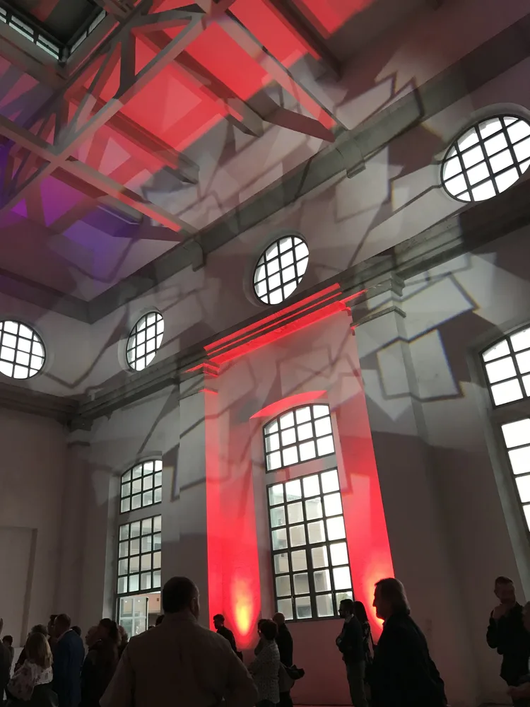 Proiezioni geometriche rosse ridisegnano pareti e soffitto della ex centrale termoelettrica di Daste e Spalenga — pubblico in piedi tra oblò e finestre industriali, composizione illuminotecnica SUONO E LUCE