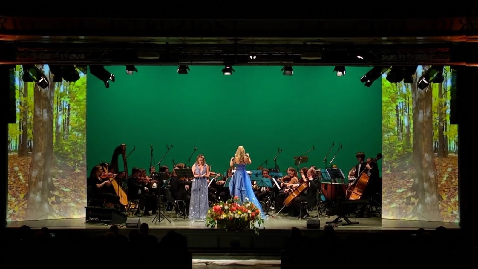Cantante solista davanti all'orchestra Ars Armonica con foresta autunnale proiettata sulle quinte — scenografia digitale, Concerto per la Natura Bergamo 2022, SUONO E LUCE