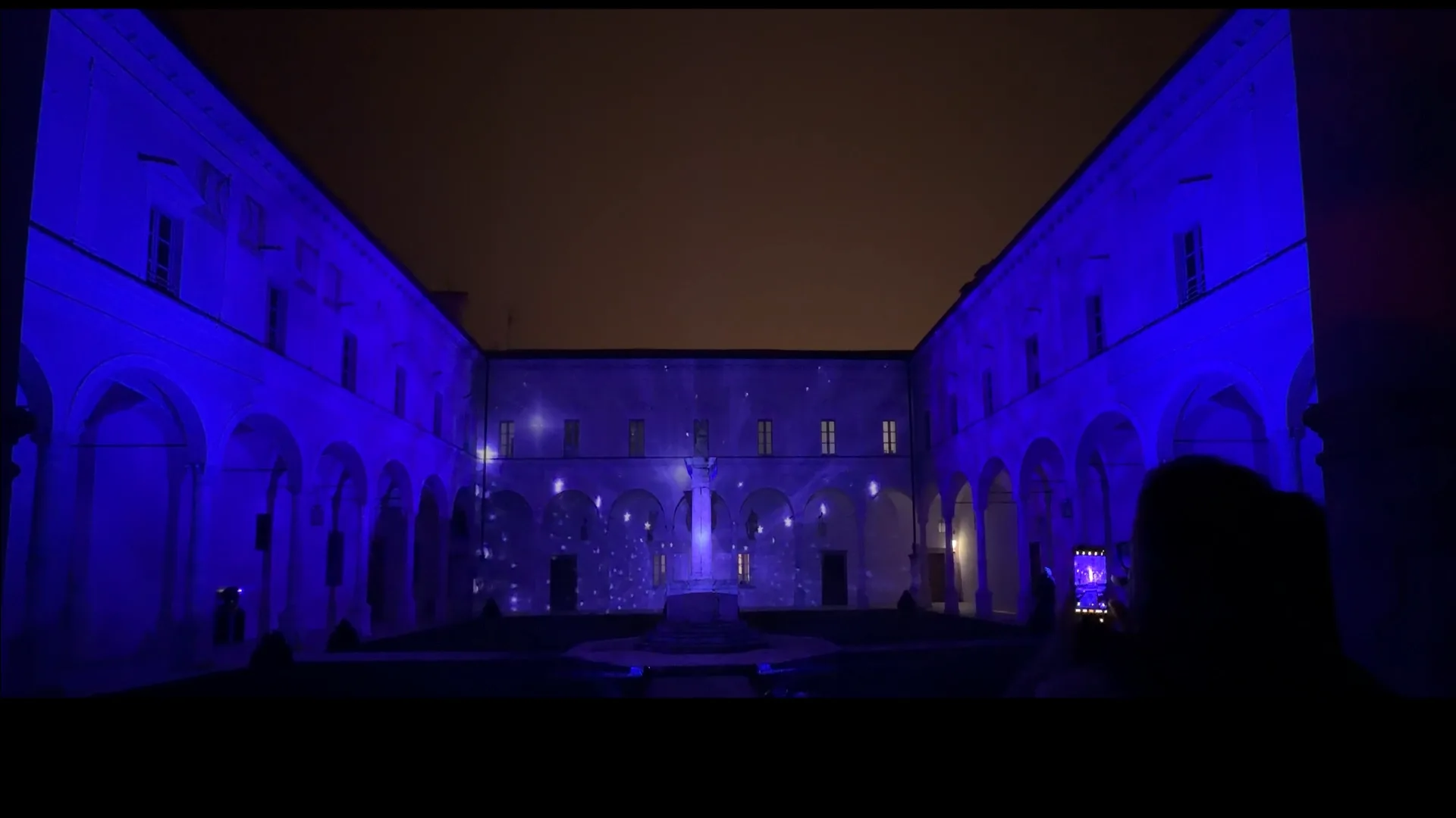 Chiostro dell'Abbazia di San Paolo d'Argon illuminato in blu con stelle e fiocchi proiettati — serata di solidarietà 19 dicembre 2025, videomapping SUONO E LUCE